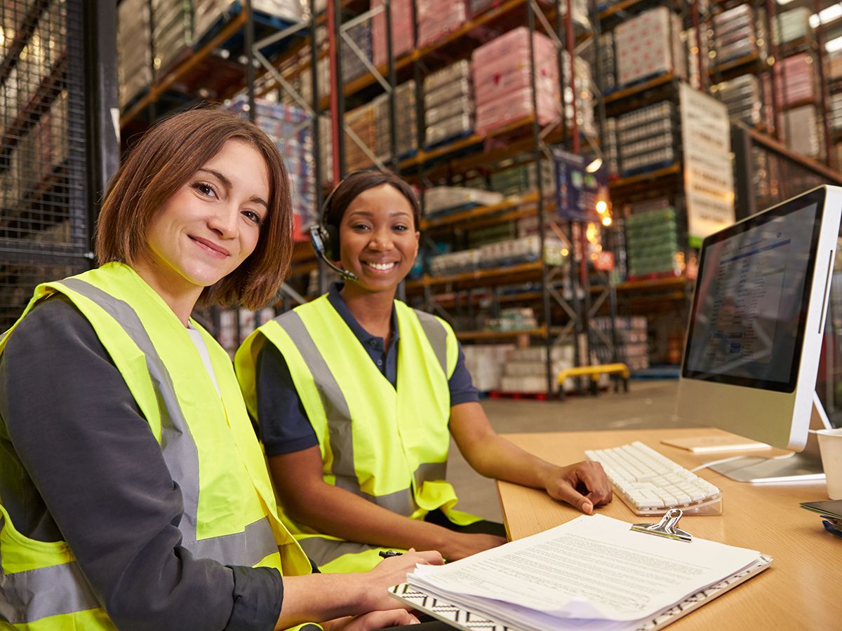 Two female colleagues in a warehouse office look to camera