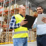 wholesale, logistic business and people concept - warehouse worker and businessman with clipboard and tablet pc computer