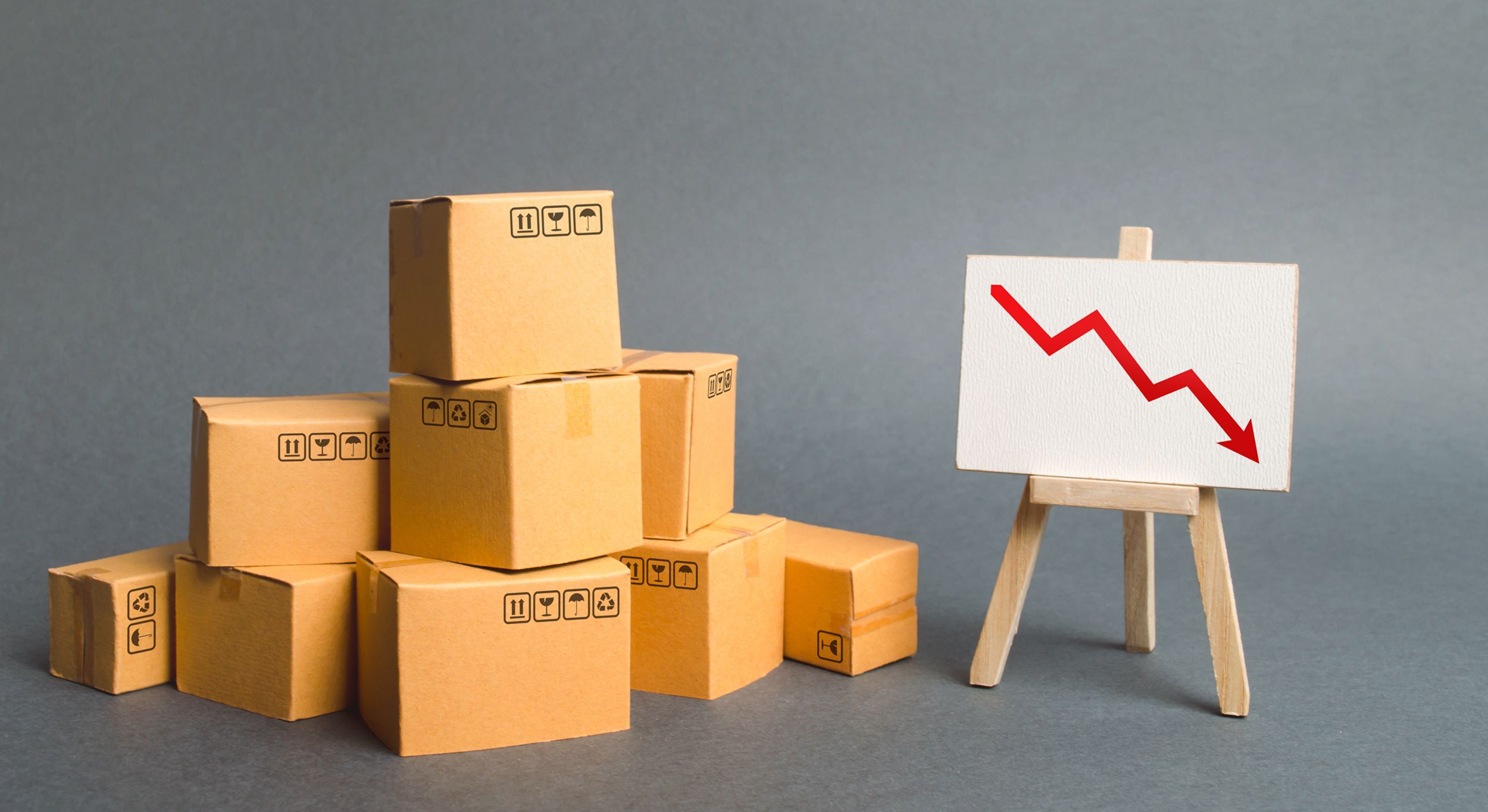 A pile of cardboard boxes and easel with red arrow down. Decrease in the quality, price, quantity and competitiveness of goods and products. Concept drop in industrial production, sales fall.
