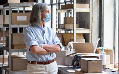 Older mature female online store small business owner, manager, stock worker, entrepreneur wearing face mask and gloves standing with arms crossed at workplace in warehouse looking through window.