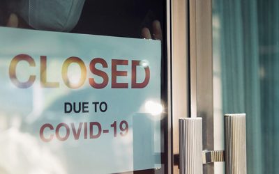 Business office or store shop is closed/bankrupt business due to the effect of novel Coronavirus (COVID-19) pandemic. Unidentified person wearing mask hanging closed sign in background on front door.